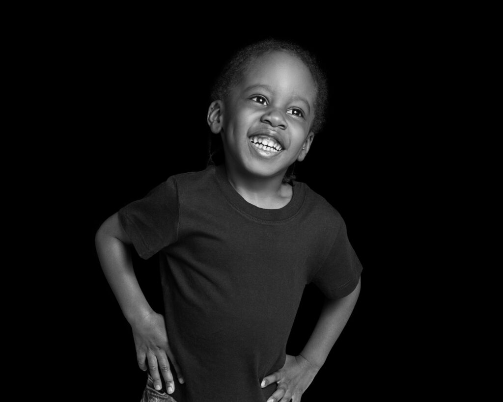 a black and white portrait of a young boy smiling with his hands on his hips. Taken at a family photograph session in Peckham, South East London.