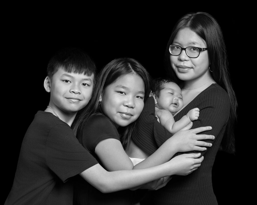 A black and white studio portrait of three siblings, holding their newborn baby sister. The photograph is elegant, natural and timeless.