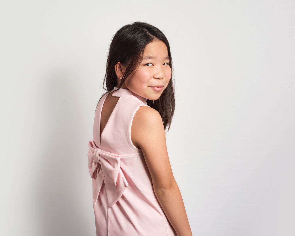 colour portrait of a young girl, looking back over her shoulder and smiling at the camera. The girl wears a pink dress and looks natural and relaxed.