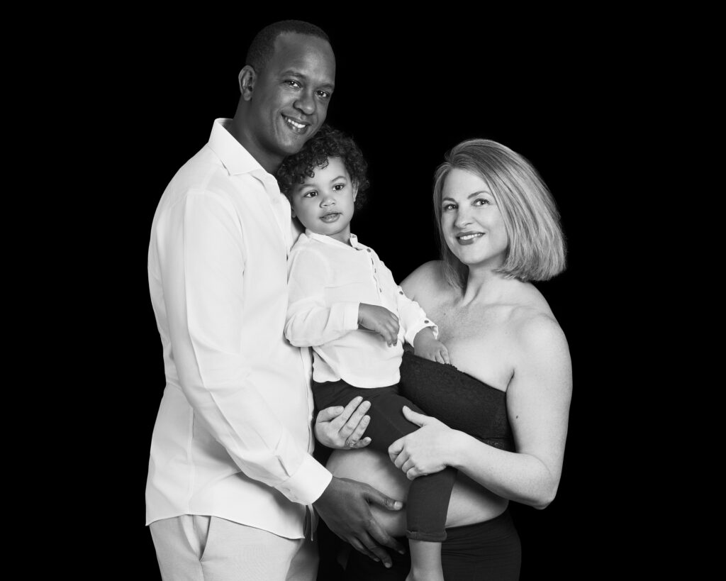 “Black and white family portrait of a pregnant mother, father, and their young son. The little boy is sitting on his mother’s tummy while his father gently touches her belly. All are smiling warmly, sharing a tender moment.