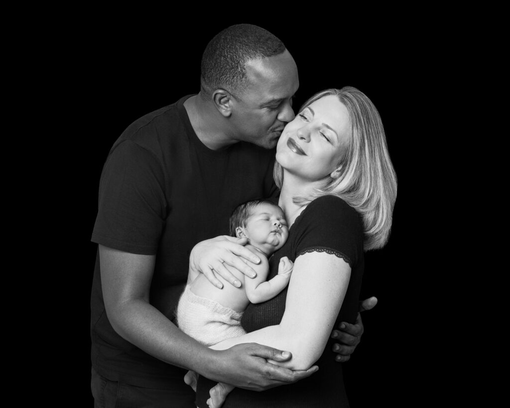 Black and white family portrait of Dad kissing Mum while she holds their newborn baby, capturing a loving and tender moment.